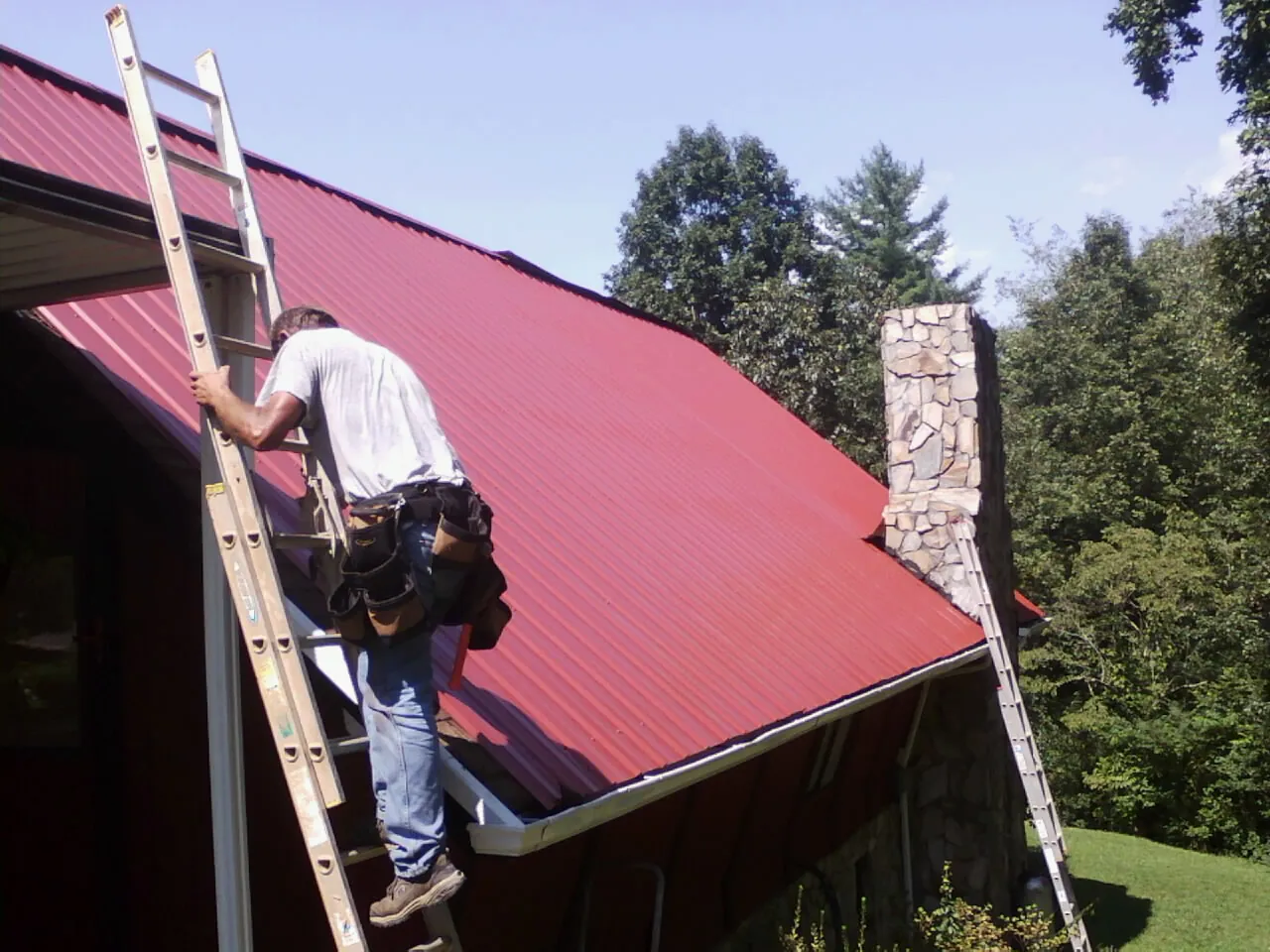 Roofer on a ladder beside a red metal roof and stone chimney — Soffit & Fascia Repair by Roofing Brothers in Augusta Springs